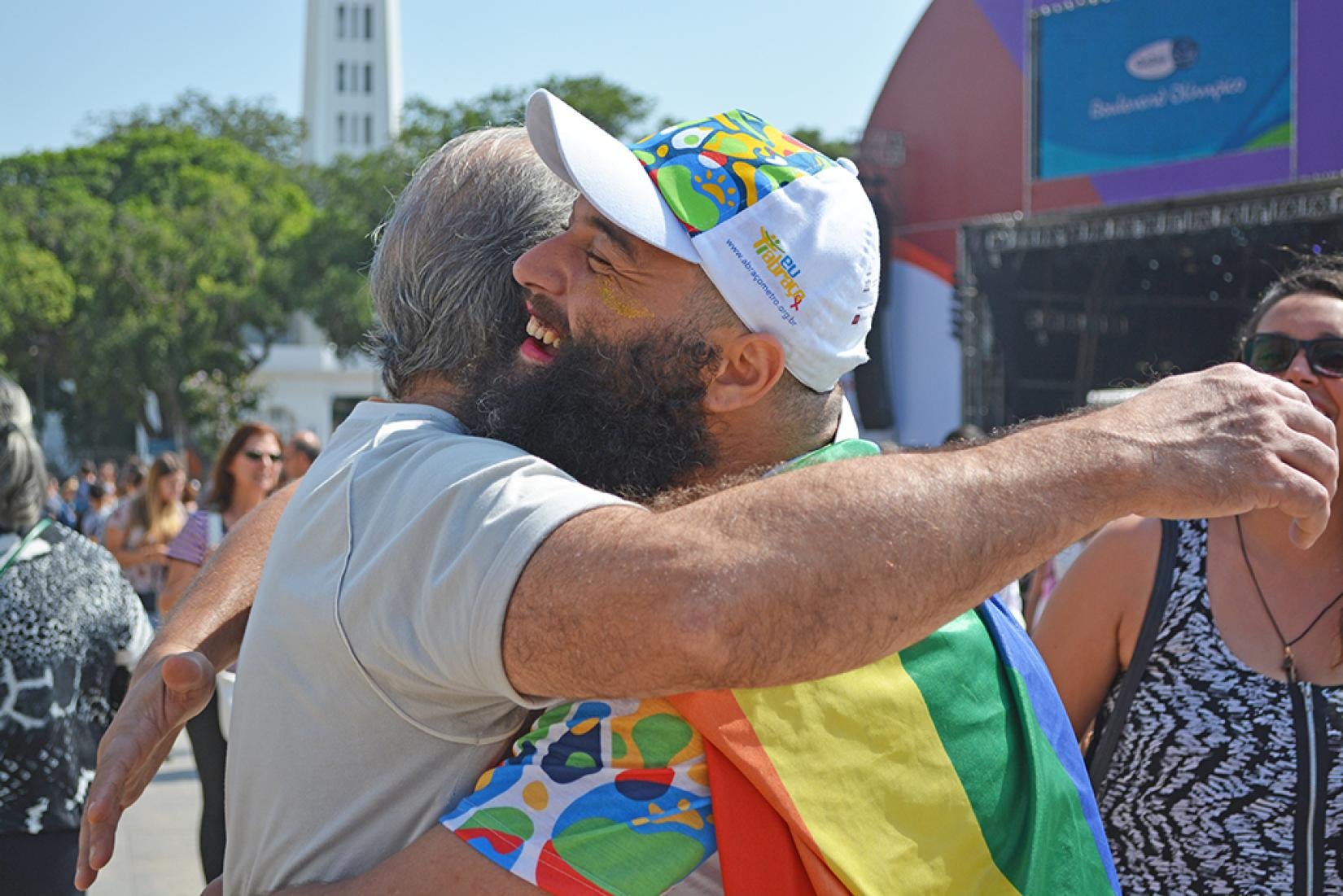 Two people hugging at the Olympic Boulevard in Rio, one wearing the campaign cap and a rainbow flag